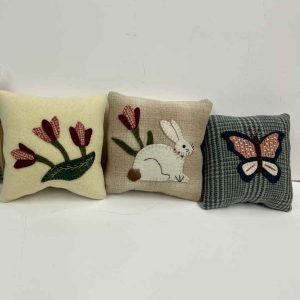 Three wool applique pin cushions - first with red tulips and green leaves on cream wool, second with white bunny and red tulips on tan wool, third with red and white butterfly on plaid wool