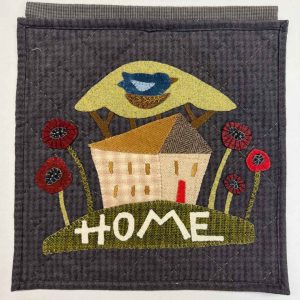 HOME wool applique wall hanging kit featuring folk art house, bluebird in yellow sun, red poppies, and primitive lettering on charcoal wool background with blanket stitching
