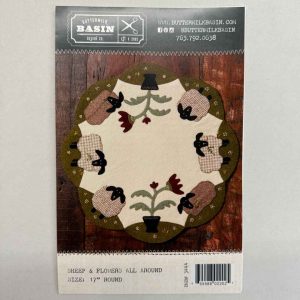 Pattern cover for Buttermilk Basin's 'Sheep and Flowers All Around' 17-inch round wool applique table mat featuring primitive sheep, black crows, burgundy flowers in pots, and scalloped olive border on cream wool background