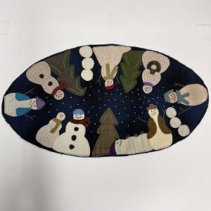 Oval wool applique piece featuring multiple snowmen arranged in a circle against a navy background with scattered snowflakes, decorated with evergreen trees and winter accessories in various textured wools