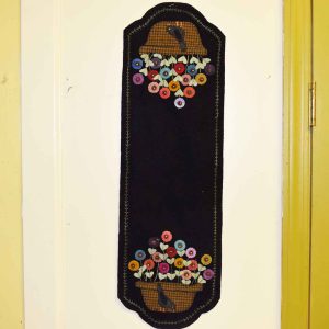 Long wool applique wall hanging on black background featuring two plaid pots with colorful penny stitched flowers and leaves, bordered with delicate green embroidery