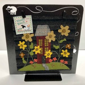 Display stand showcasing a wool applique design titled 'Wooly Block 2018', featuring a red saltbox house with gray roof surrounded by yellow, tan and orange flowers on a dark background, with a decorative sheep silhouette at top