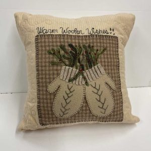 Wool applique holiday pillow with 'Warm Woolen Wishes' text, featuring tan mittens embroidered with chevron patterns and evergreen sprigs with red berries on brown plaid background
