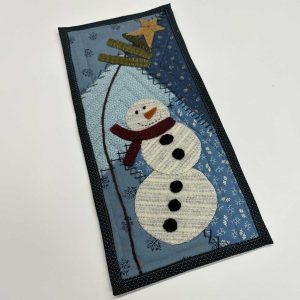 Quilted winter wall hanging featuring an appliqued snowman with a burgundy scarf against a blue background with snowflakes, topped with a gold fabric star and evergreen branches