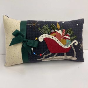 Christmas wool applique pillow featuring a red sleigh filled with presents and greenery on navy background with embroidered stars, decorated with green grosgrain bow and cream polka dot fabric accent