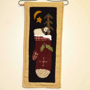 Wool applique wall hanging with red mitten on black background bordered in tan plaid, featuring a sleeping lamb, embroidered snowflake, evergreen tree with red berries, and gold star and moon motifs