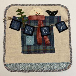 Winter wool applique wall hanging kit featuring snowman wearing plaid scarf, SNOW banner, evergreen tree, and black bird on cream background with blue floral border and striped binding