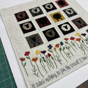 Wool applique hanging featuring twelve squares with sheep silhouettes in various fabrics, bordered by embroidered wildflowers at the bottom and text reading 'It takes nothing to join the crowd...'